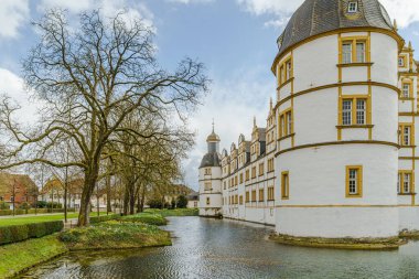 Paderborn Almanya 'sında sarı çatılı ve saat kulesi olan Neuhaus Kalesi' nin beyaz bir binası. Bina ağaçlar ve bir gölet ile çevrili..