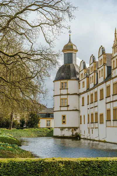 Paderborn Almanya 'sında Neuhaus Kalesi' nin önünde bir kule ve gölet olan bir bina. Gölet sakin ve bina beyaz. Fotoğraf dikey biçimdeName.