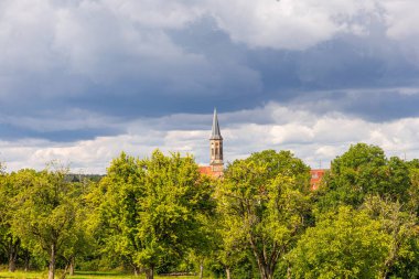 Almanya 'nın arka planında yüksek bir kilise binası olan bulutlu bir gökyüzü. Ağaçlar yeşil ve çimenler gür..