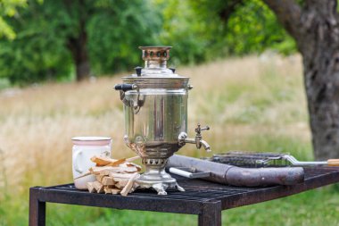 Semaver, dışarıda, ormandaki bir açıklıkta demir bir masanın üzerinde duruyor. Semaverin yanında doğranmış odun ve diş minesi çaydanlığı var. Boru da görünür durumda..