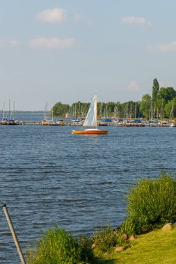 Üzerinde birkaç teknenin yüzdüğü sakin bir göl. Tekneler çeşitli boylarda ve renklerde ve gölün ortasında bir yelkenli var. Huzurlu ve huzurlu bir sahne..