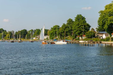 Üzerinde birkaç teknenin yüzdüğü sakin bir göl. Tekneler çeşitli boylarda ve renklerde ve gölün ortasında bir yelkenli var. Huzurlu ve huzurlu bir sahne..