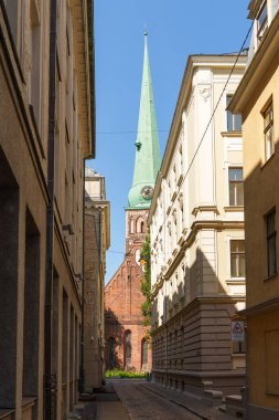 Letonya 'nın Riga şehrinde arka planda bir kilisenin olduğu dar bir sokak. Kilise uzun ve yeşildir. Gökyüzü mavi ve berrak.