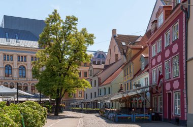 Riga Letonya 'daki güneş ışığı parke taşı caddesi renkli tarihi binalarla doludur. Pembe ve beyaz cepheler arka plandaki modern çatılarla tezat oluşturuyor. Büyük ağaç, açık kafe koltuklarına gölge veriyor..