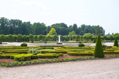 Manikürlü çitleri ve çeşme süsü olan simetrik bahçe. Yeşillik, berrak gökyüzü altında durgun su özelliğini çerçeveler. Uzun ağaçlar geometrik tasarımlara zıt arkaplan sunar.