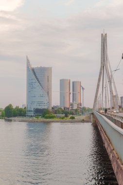Riga Letonya 'da geniş bir köprünün yanında modern kuleler yükseliyor. Gökyüzü yumuşaktır, bulutlar yavaşça yukarı doğru sürüklenir. Su, cam ve çeliği zarafetle yansıtır. Kentsel ufuk çizgisi açık gökyüzü ile durgun denge içinde buluşuyor.
