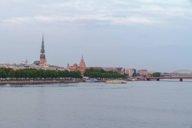 Günbatımı Riga Letonya 'da eski kasabayı sıcak, altın bir ışıkla yıkıyor. Duna Nehri, tarihi binaların ve ağaçların yanında nazikçe akıyor. Kuleler ve çatılar, sakin ufuk çizgisini çerçeveliyor..