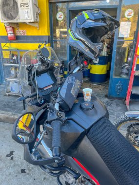 Macera motosikletli gösterge panelinin önünde kağıt kahve fincanı ve benzin istasyonunun gidonunda miğfer var..