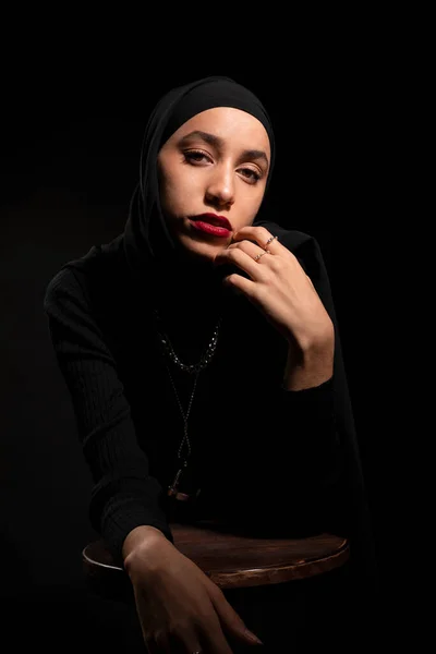 魅力的な若いですイスラム女性身に着けています黒服とヒジャーブのタッチ顔優しく黒スタジオで椅子にもたれてカメラを見て — ストック写真