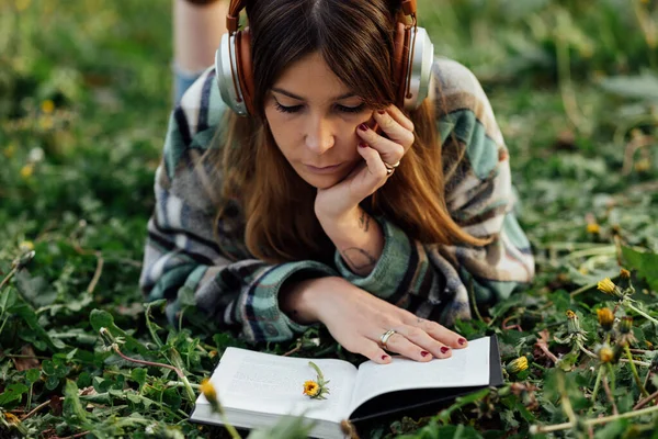 Young Attentive Female Modern Headset Reading Textbook While Lying Meadow — Fotografia de Stock