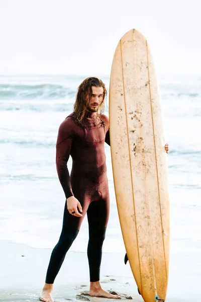 長い髪をした若いサーファーの男は 日の出の間にビーチで波をキャッチするために水に向かってサーフボードとカメラを見て立っているウェットスーツに身を包んだ — ストック写真
