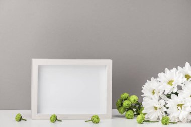 Boş fotoğraf çerçevesi, beyaz kasımpatılar, gri bir duvara karşı beyaz bir masada papatyalar. Kartpostal, sertifika, ev içi.