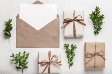 Beyaz arka planda üç hediye kutusu, yeşil boxwood dalları ve içinde boş bir kart olan bir el işi zarfı. El yapımı hediyeler. Kartpostal, maket.