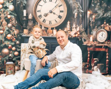 Baba ve oğul Noel süslemeli odada gülümsüyor. Büyük, eski bir saat ve hediyelerle. Sıcak aile havası