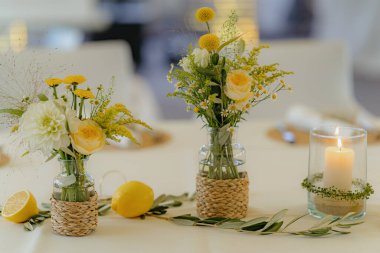 Örülmüş vazolarda taze sarı ve beyaz çiçekler, limonlar ve cam tutacağında yanan bir mum bulunan güzel masa süsü. Düğünler, etkinlikler ya da ev dekorasyonu için mükemmel