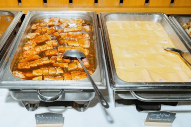 Büfede soslu dilimlenmiş domuz eti tepsileri ve kremalı soslu morina filetosu var. Her tepside bir metal kepçe var, kişisel servis ayarlarını vurguluyor.