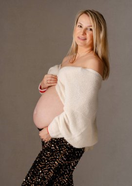 28 hafta, krem örgü örmüş, gülümseyen bir annenin sıcak stüdyo portresi, elleri bebek karnında dinleniyor, neşeli ve samimi bir şekilde hamilelik parıltısını yakalıyor.