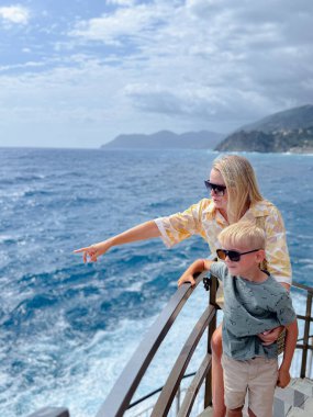 Anne ve oğlu kıyı korkuluklarında birlikte manzaranın tadını çıkarıyorlar. Anne hem güneş gözlüğü takıyor hem de mutluluk ve memnuniyet sergiliyor..