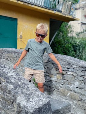 Genç bir çocuk, canlı sarı bir binanın yakınındaki açık havada, taş bir duvarda güven içinde dengede duruyor. Güneş gözlüğü ve günlük kıyafetlerle, macera ve oyun anlayışı yayıyor..