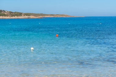 Şeffaf turkuaz suyun kumlu sahilde yavaşça yıkandığı sakin bir sahil sahnesi. Parlak ufuk ve ılık gün ışığı huzurlu bir tatil ortamı yaratır. Seyahat görüntüsü için mükemmel, La