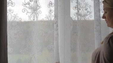 Yumuşak gün ışığı, narin dantel perdeye uzanan kadının elini vurgular. Sahne, sakin bir düşünce anını yakalar. Yumuşak dokular, sıcak tonlar ve huzurlu bir ortam.