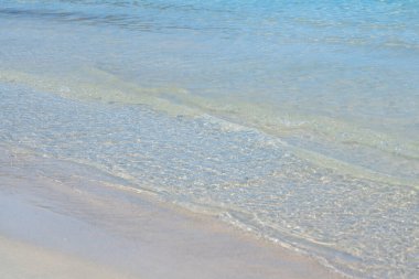 Yumuşak dalgalar sığ kumlu bir kıyı şeridinde süzülerek turkuaz deniz suyunun berraklığını açığa çıkarıyor. Yumuşak doku ve doğal ışık huzurlu bir sahil atmosferini taşır.