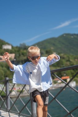 Neşeli bir çocuk, dağlarla çevrili bir göl kenarındaki korkuluklara yakın poz veriyor ve parlak yaz ışığı. Neşeli jest ve şık güneş gözlükleri enerjik bir açık hava portresi oluşturur.
