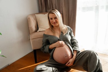 34 hafta, hamile bir kadının gün ışığında koltuğun yanında oturduğu nazik bir doğum anı. Sakin tonlar ve rahat duruş samimi ve sıcak bir yaşam tarzı görüntüsü oluşturur