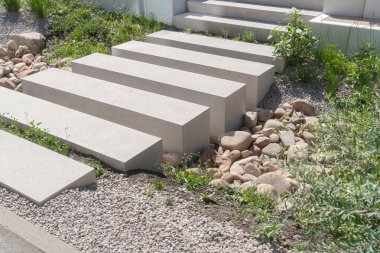 Çakıl ve yeşillik arasında sıralanmış beton basamakları olan modern bir bahçe yolu. Temiz geometrik düzen ve doğal dokular, minimal ve çağdaş görünümlü dengeli bir açık hava manzarası yaratır.