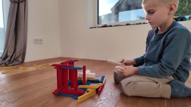 Bir çocuk yere oturur ve renkli ahşap parçaları küçük bir yapıya yerleştirir. Parlak kapalı mekan sahnesi erken öğrenmeyi, yaratıcılığı ve odaklanmış oyunu ifade eder.