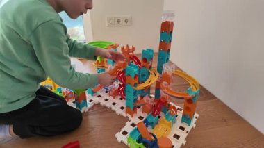 Genç bir çocuk yerde oturuyor ve renkli bir mermer set ile etkileşime geçiyor. Parlak plastik yapılar merakı, yaratıcılığı ve erken mühendislik keşiflerini vurgular