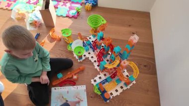 Bir çocuk rampalar, huniler ve renkli plastik elementlerin yer aldığı geniş bir mermer koşu takımının yanında oturuyor. Sahne, eğlenceli keşifler, yaratıcılık ve erken dönem STEM tarzı öğrenme içeriyor.