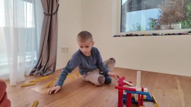 Küçük bir çocuk ahşap zeminde oturuyor ve kırmızı ve mavi ahşap bir yapıyla etkileşime giriyor. İçerideki sakin ortam yaratıcılığı, iyi motor becerileri ve basit yaratıcı oyunu ön plana çıkarıyor.