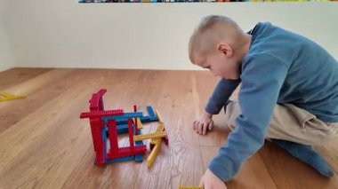 Bir çocuk ahşap bir zeminde oturuyor ve kırmızı ve mavi bir tahta blok yapısıyla etkileşime giriyor. Sahne yaratıcılığı, odaklanmayı ve basit hayal gücünü gösteriyor. Parlak ve samimi kapalı bir ortamda..