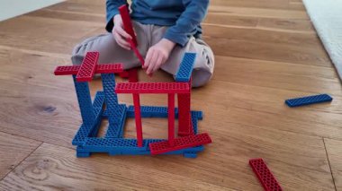 Bir çocuk yerde otururken kırmızı ve mavi bir ahşap blok yapısını birleştirir. Sahne, yaratıcılığı, odaklanmayı ve yapıcı oyunu kapalı alan ortamında yakalıyor.
