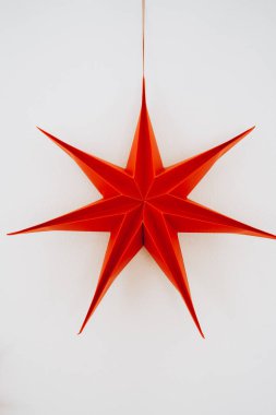 Temiz beyaz bir duvarda gün ışığında asılı duran büyük kırmızı kağıt yıldız süsü. Basit ve çarpıcı Noel dekoru modern iç mekanlar, şenlikli tasarımlar ve mevsimsel tasarımlar için mükemmeldir.