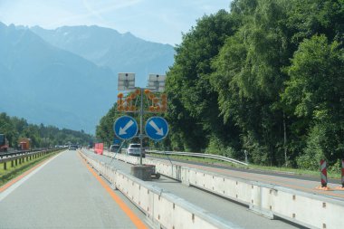 Arkasında beton bariyerler, tabelalar ve dağlar olan bir otoyol bölümü. Doğal yeşillik ve yaz ışığı karşıtlığı, altyapı için uygun çalışma alanı ve