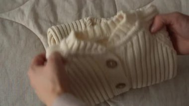 Bir kadın eli, yumuşak örülmüş bebek kıyafetlerini düzgünce yatağa katlar. Ilık doğal ışık, kumaşın dokusunu ön plana çıkarır. Yeni doğan annelik temaları için ideal yumuşak ve rahat bir atmosfer yaratır.