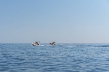 Sakin bir açık deniz, açık bir gökyüzü altında hafif dalgalar. Yumuşak gün ışığı su yüzeyine yansıyarak huzurlu bir deniz manzarası yaratır. Uzakta, iki küçük motorlu tekne ve tatilciler karşıya geçiyor.