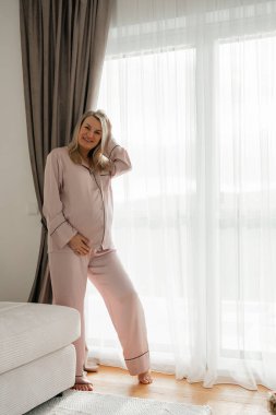 36 hafta, Pastel giyim giymiş bir kadın büyük bir pencerenin yanında, parlak bir oturma odasında duruyor. Yumuşak doğal ışık, sakin ve havadar bir atmosfer yaratır. Modern iç mekan ayrıntıları rahat yaşam tarzını tamamlar