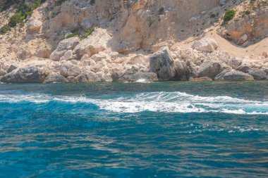 Küçük dalgalar gün ışığında kayalık kıyı şeridine hafifçe çarpıyor. Kalıplaşmış kaya kayalıkları ve derin mavi su hareket ve enerji dolu doğal bir kıyı sahnesi oluşturur.