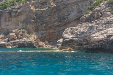 Engebeli bir kayalık kıyı şeridi berrak bir yaz gökyüzü altında durgun turkuaz suyun üzerinde yükseliyor. Sıcak gün ışığı taş dokuları vurgular, temiz ve doğal bir Akdeniz manzarası yaratır