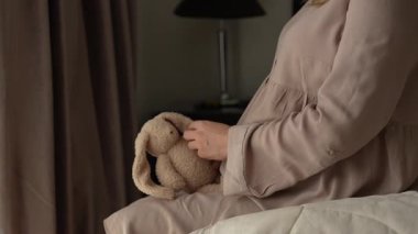 Bebek oyuncağı tutan hamile bir kadın yatakta oturuyor. Sakin bir ev, annelik ve doğum için duygusal hazırlık.