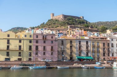 Rıhtım boyunca uzanan renkli binaların üzerinde tarihi bir tepe kalesi olan bir Avrupa kasabası. Parlak gün ışığı, mimari zıtlıkları ve kıyı cazibesini vurguluyor, Bosa, Italien