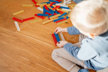 Küçük bir çocuk ahşap bir zeminde renkli yapı taşlarından yüksek bir yapı oluşturuyor. Odaklanmış oyun ve parlak elementler çocukluk gelişimini, yaratıcılığı, problem çözmeyi ve hayal gücünü ön plana çıkarıyor