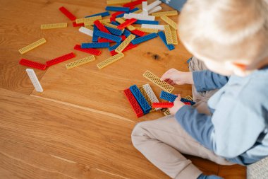 Ahşap bir zeminde oturan çocuk renkli bina oyuncaklarıyla çevrili, taşları tutan ve yaratıcı oyunlarla meşgul. Sıcak ev atmosferi, erken gelişim, hayal gücü ve