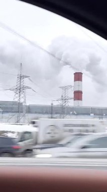 Endüstriyel binalar ve uzun bacalar buhar salıyor, bir arabanın camından çekiliyor. Trafik, elektrik hatları ve kentsel yaşamı yansıtan soğuk renklerle dinamik bir kış şehri.