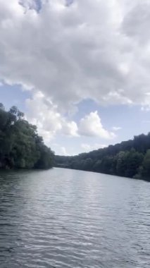 Bulutlu bir gökyüzünün altında sık ormanlarla çevrili sakin bir nehrin huzurlu manzarası. Yumuşak su hareketleri ve doğal manzara doğa temaları, seyahat, meditasyon ve diğer şeyler için uygun rahatlatıcı bir atmosfer yaratır.