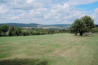 Parlak, bulutlu bir gökyüzünün altında yeşil tarlaları ve uzak tepeleri olan geniş bir kırsal alan. Ufuk boyunca uzanan yemyeşil ağaçlar, sakin ve görkemli bir manzara yaratıyor..