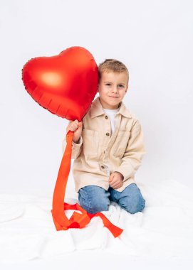 Küçük çocuk oturuyor ve kırmızı kalp balonunu suratının önünde tutuyor. Sevgililer Günü, neşeli ruh hali, aşk, çocukluk masumiyeti.